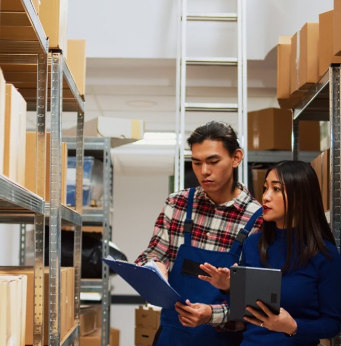 Asian employees checking warehouse supplies on tablet and files, working with storage room logistics. Young team of people using gadget and papers for business plan and production.