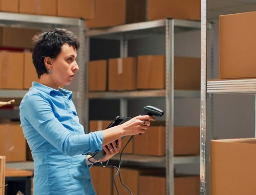 Business owner scanning stock from storehouse shelves, working with scanner to do inventory and check logistics supplies. Female employee using tablet to scan bar code on merchandise. Handheld shot.
